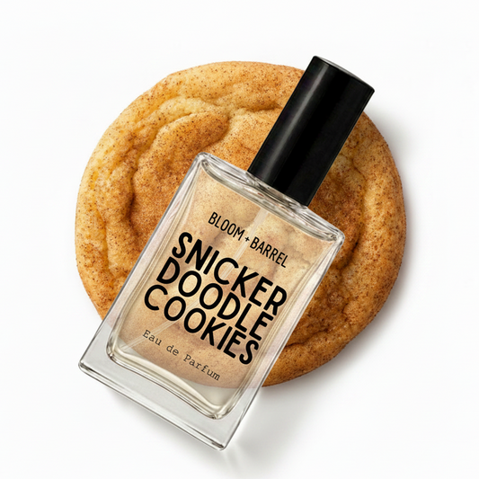 A perfume bottle labeled 'BLOOM • BARREL' and 'SNICKER DOODLE COOKIES' stands in front of a large sugar-coated cookie on a white background.