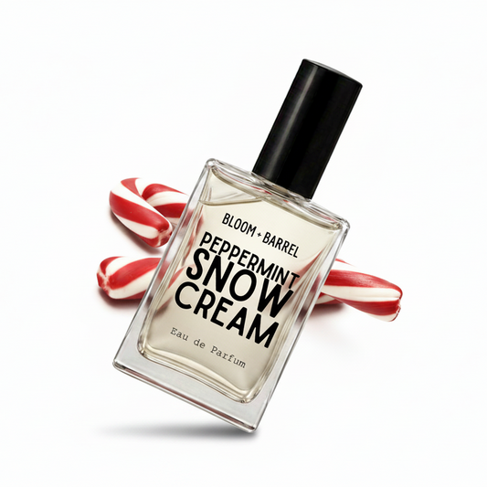 A clear glass perfume bottle labeled 'BLOOM • BARREL PEPPERMINT SNOW CREAM Eau de Parfum' with a black cap, centered on a white background and flanked by two red-and-white striped candy canes.