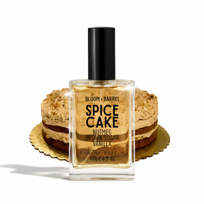 A bottle of Bloom + Barrel Spice Cake Eau de Parfum centered in front of two layered brown sugar and vanilla cake slices on gold platters, against a plain white background.