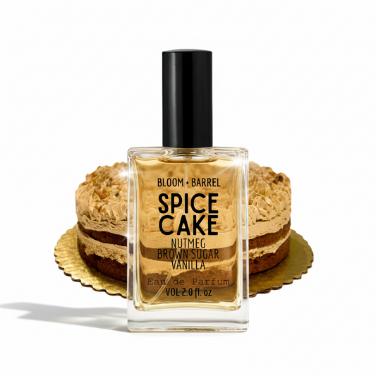 A bottle of Bloom + Barrel Spice Cake Eau de Parfum centered in front of two layered brown sugar and vanilla cake slices on gold platters, against a plain white background.