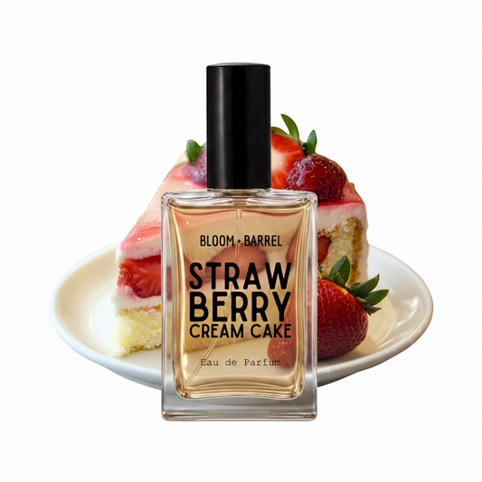 A perfume bottle labeled 'BLOOM • BARREL STRAW BERRY CREAM CAKE' stands in front of a sliced strawberry cream cake on a white plate against a plain white background.