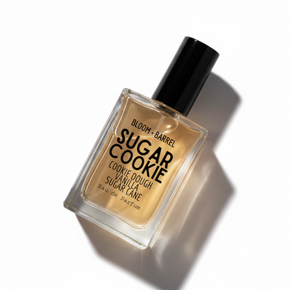 A rectangular glass perfume bottle with a black cap, filled with amber-colored fragrance liquid. The front label reads 'BLOOM • BARREL SUGAR COOKIE COOKIE DOUGH VANILLA SUGAR CÂNE Eau de Parfum' in black text, set against a plain white background.