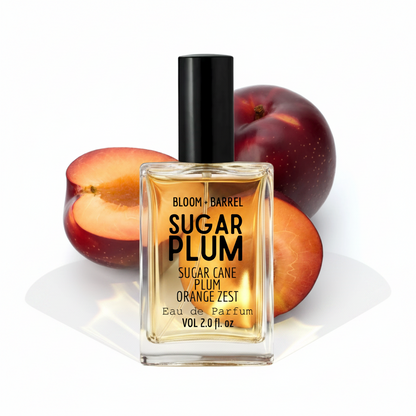 A bottle of Bloom + Barrel Sugar Plum Eau de Parfum with a black cap and clear glass, set against sliced and whole plums on a white background.