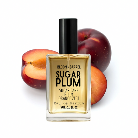 A bottle of Bloom + Barrel Sugar Plum Eau de Parfum with a black cap and clear glass, set against sliced and whole plums on a white background.