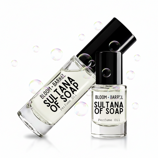 Two clear glass perfume oil bottles with black caps from BLOOM + BARREL, labeled 'SULTANA OF SOAP', shown on a white background with floating iridescent bubbles.