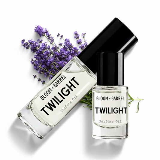 Two clear glass perfume oil bottles with black caps from Bloom + Barrel, labeled Twilight, arranged on a white background with purple lavender flowers and green stems as props.