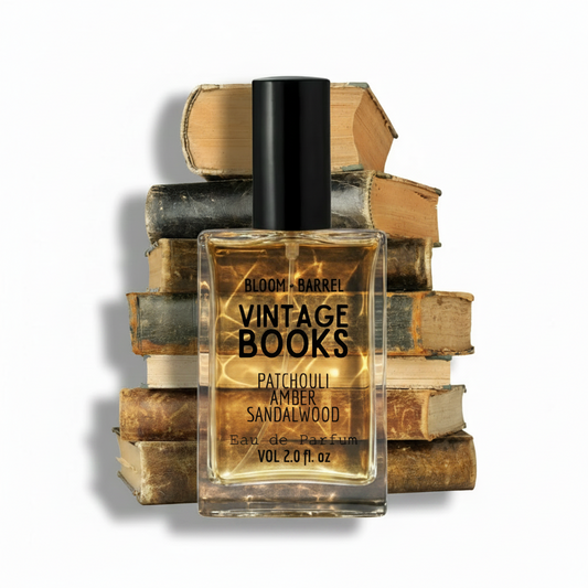 A rectangular glass perfume bottle labeled 'BLOOM + BARREL Vintage Books Patchouli Amber Sandalwood Eau de Parfum' stands centered in front of a stacked pile of old books on a plain white background.