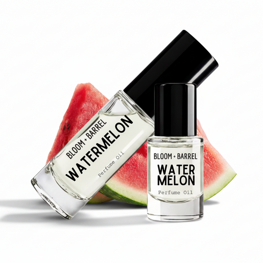 Two clear glass perfume oil bottles with black caps labeled BLOOM + BARREL WATERMelON, shown in front of watermelon slices on a white background.