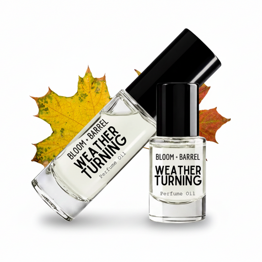 Two glass bottles of Bloom + Barrel Weather Turning perfume oil and perfume oil with black caps, styled with yellow and orange autumn leaves on a white background.