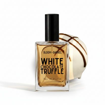 A rectangular glass perfume bottle with a black cap, filled with amber liquid, labeled 'BLOOM • BARREL WHITE CHOCOLATE TRUFFLE Eau de Parfum.' A white chocolate truffle with dark drizzle sits behind the bottle on a clean white background.