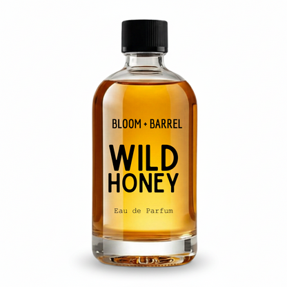 A clear glass bottle of Bloom + Barrel Wild Honey Eau de Parfum with amber-colored liquid, a black cap, and bold black text on a white background.