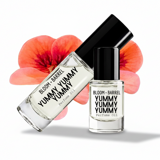 Two bottles of Bloom + Barrel Yummy Yummy Perfume Oil with black caps and clear glass, shown on a white background with coral flower graphics behind them.